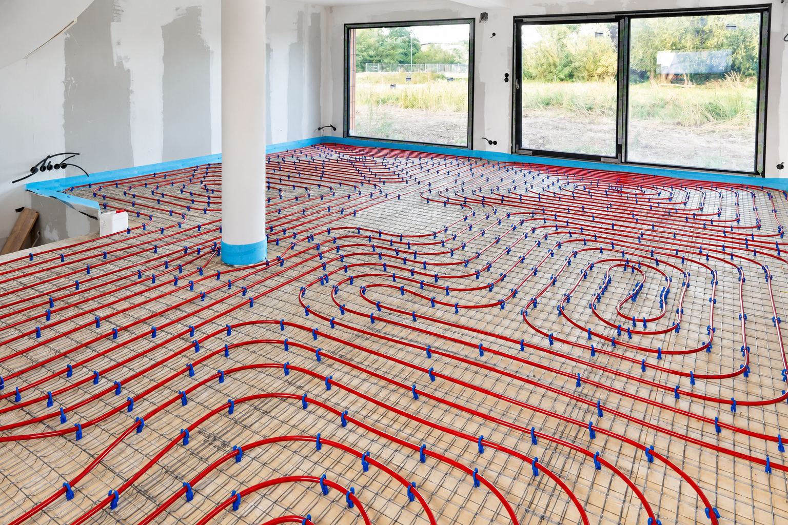 Des tuyaux rouges de chauffage au sol sont posés en boucles sur un treillis métallique dans une pièce en travaux avec de grandes baies vitrées.