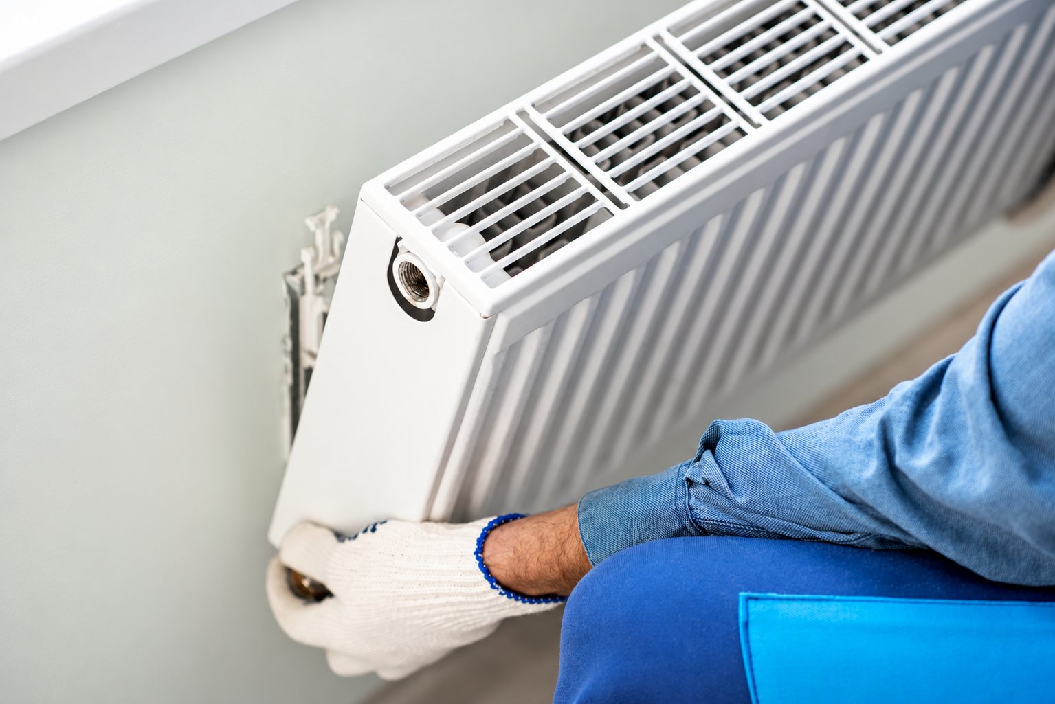 Un installateur en vêtements bleus et gants de travail met en place un radiateur à panneau blanc contre un mur clair.