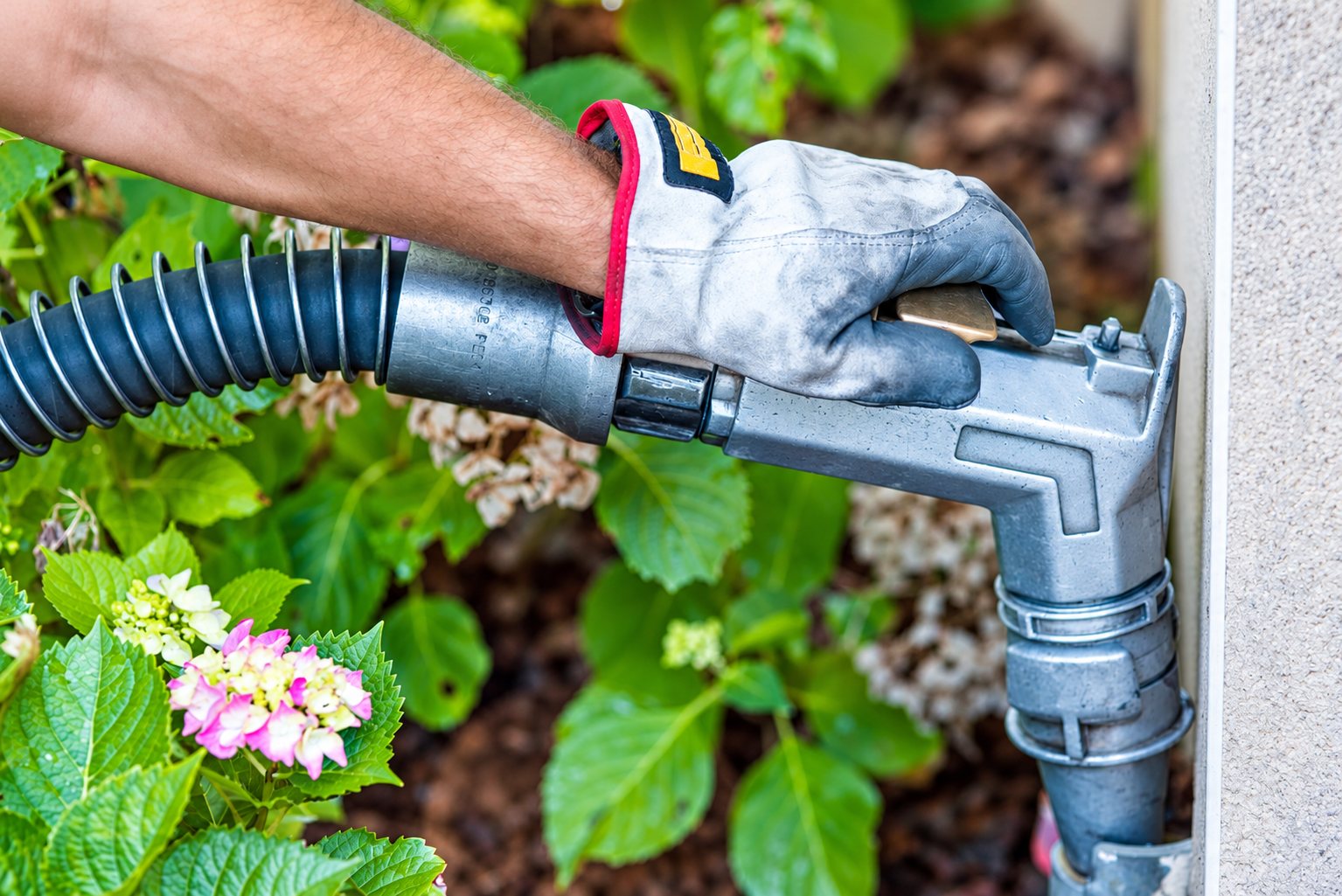 Une main gantée tient un pistolet de remplissage relié à un tuyau flexible pour alimenter une cuve de fioul, au milieu de feuillages et d'hortensias roses.