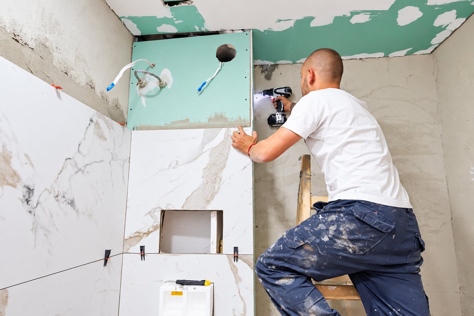 Un ouvrier en t-shirt blanc, perché sur une échelle, utilise une perceuse sans fil sur une cloison en plaque de plâtre verte dans une salle de bain en cours de rénovation avec du carrelage effet marbre.