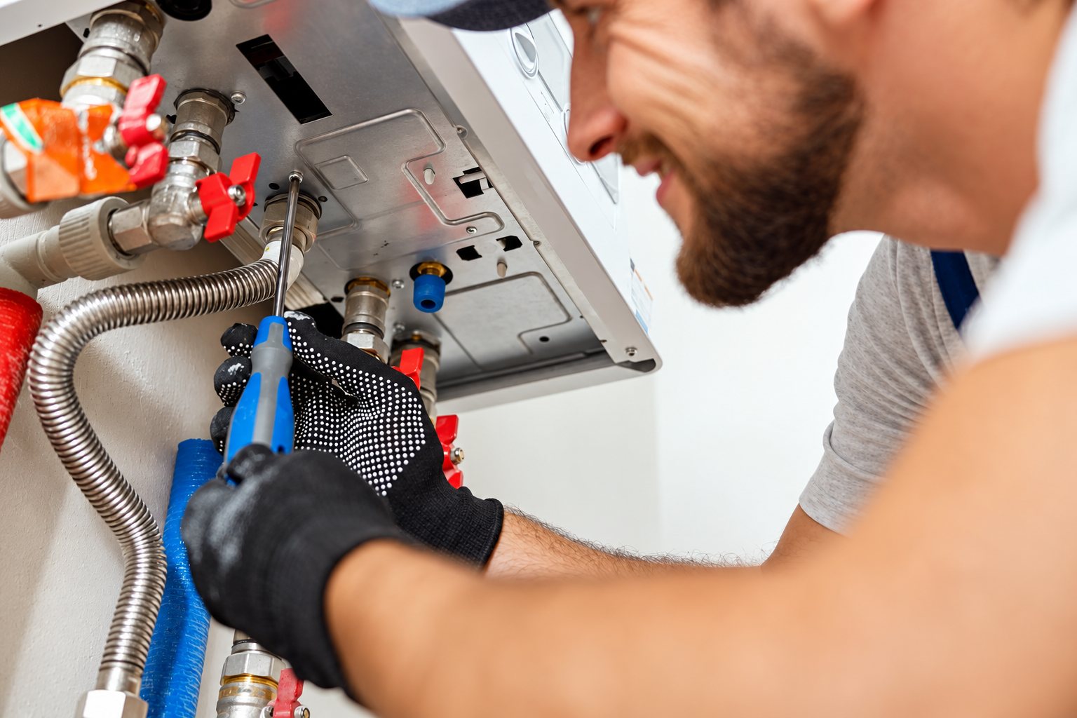 Un plombier barbu souriant, portant des gants, utilise un tournevis pour intervenir sur les raccords d'une chaudière murale.