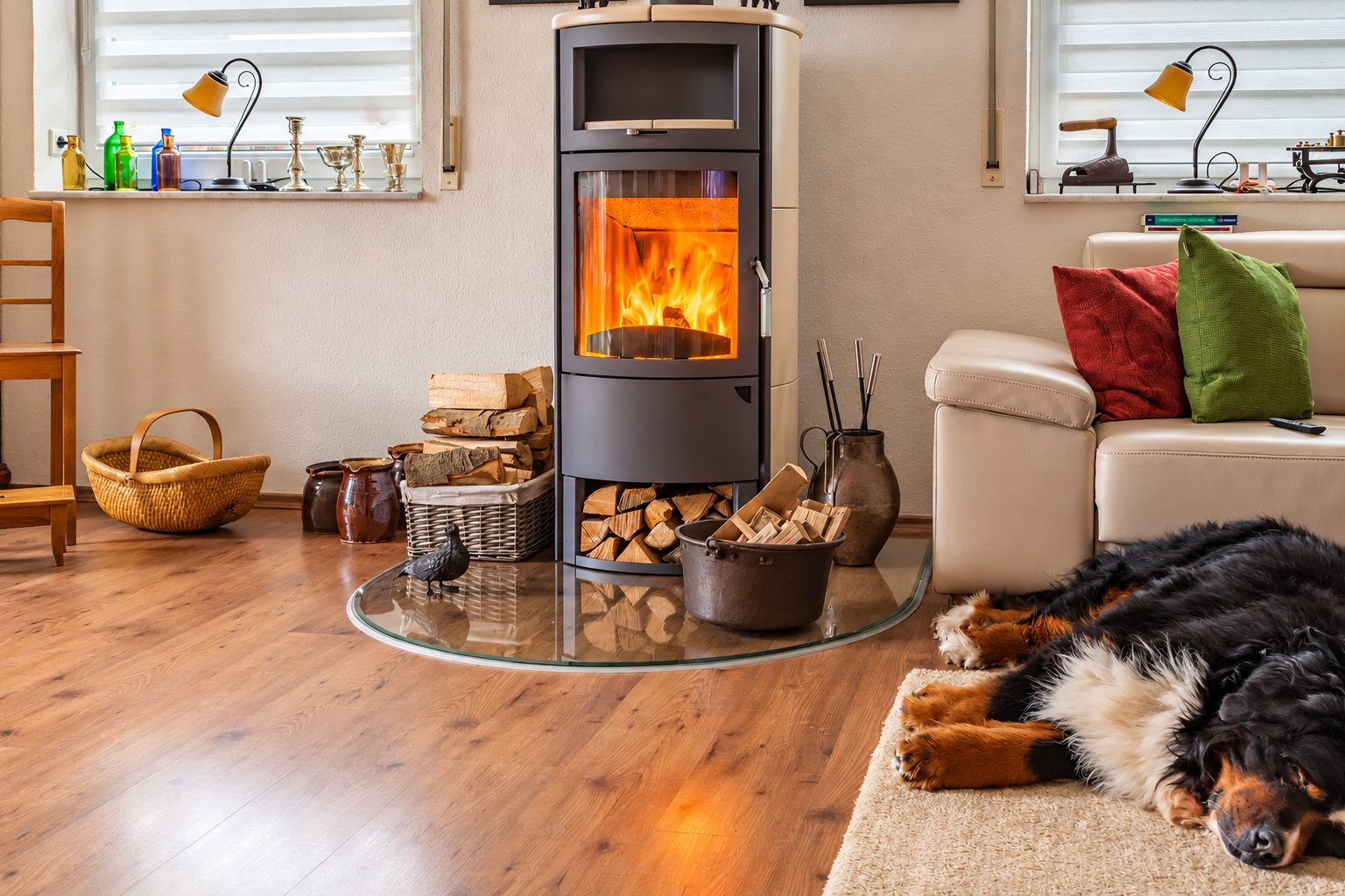 Un poêle à bois cylindrique gris avec flammes visibles trône sur une plaque de verre dans un salon chaleureux, tandis qu'un bouvier bernois dort sur un tapis près du canapé.