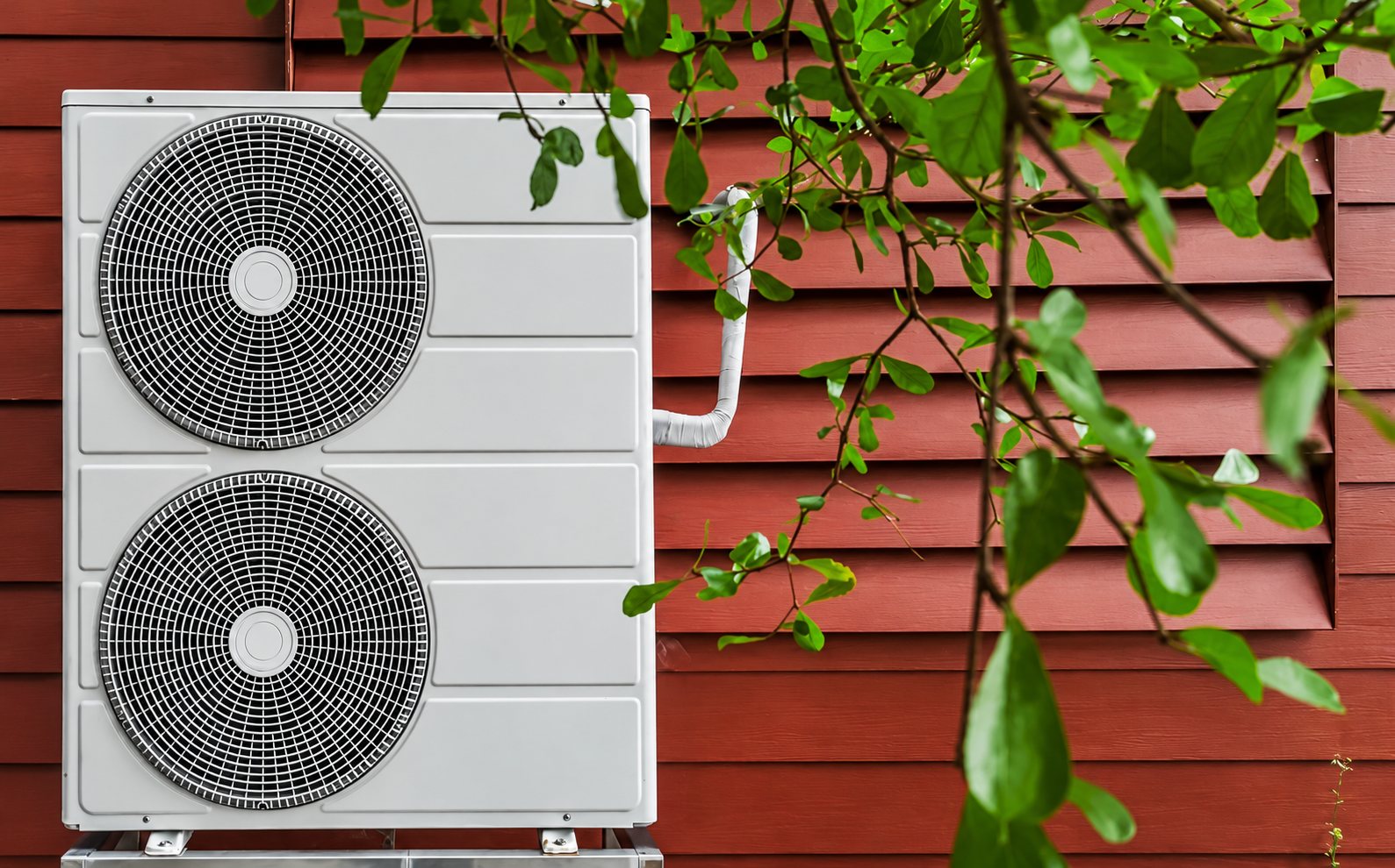 Une unité extérieure de pompe à chaleur à deux ventilateurs est fixée devant un bardage en bois rouge, avec des feuilles vertes au premier plan.