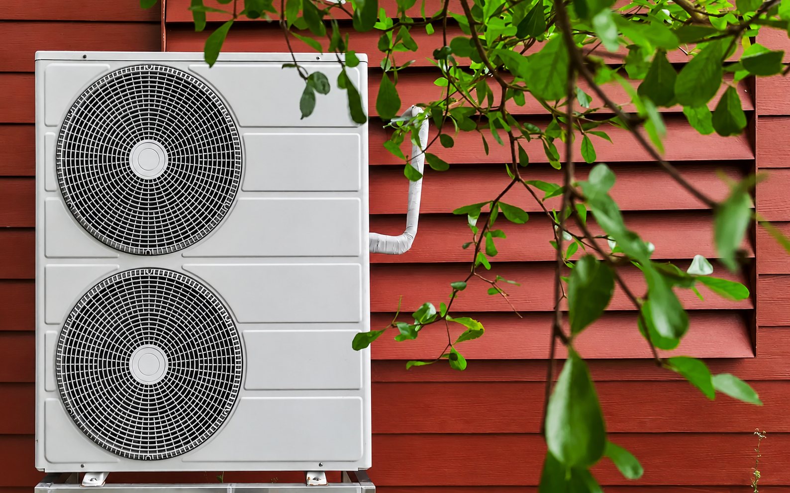Une unité extérieure de pompe à chaleur blanche à deux ventilateurs est fixée devant un mur en bardage rouge, avec des feuilles vertes au premier plan.