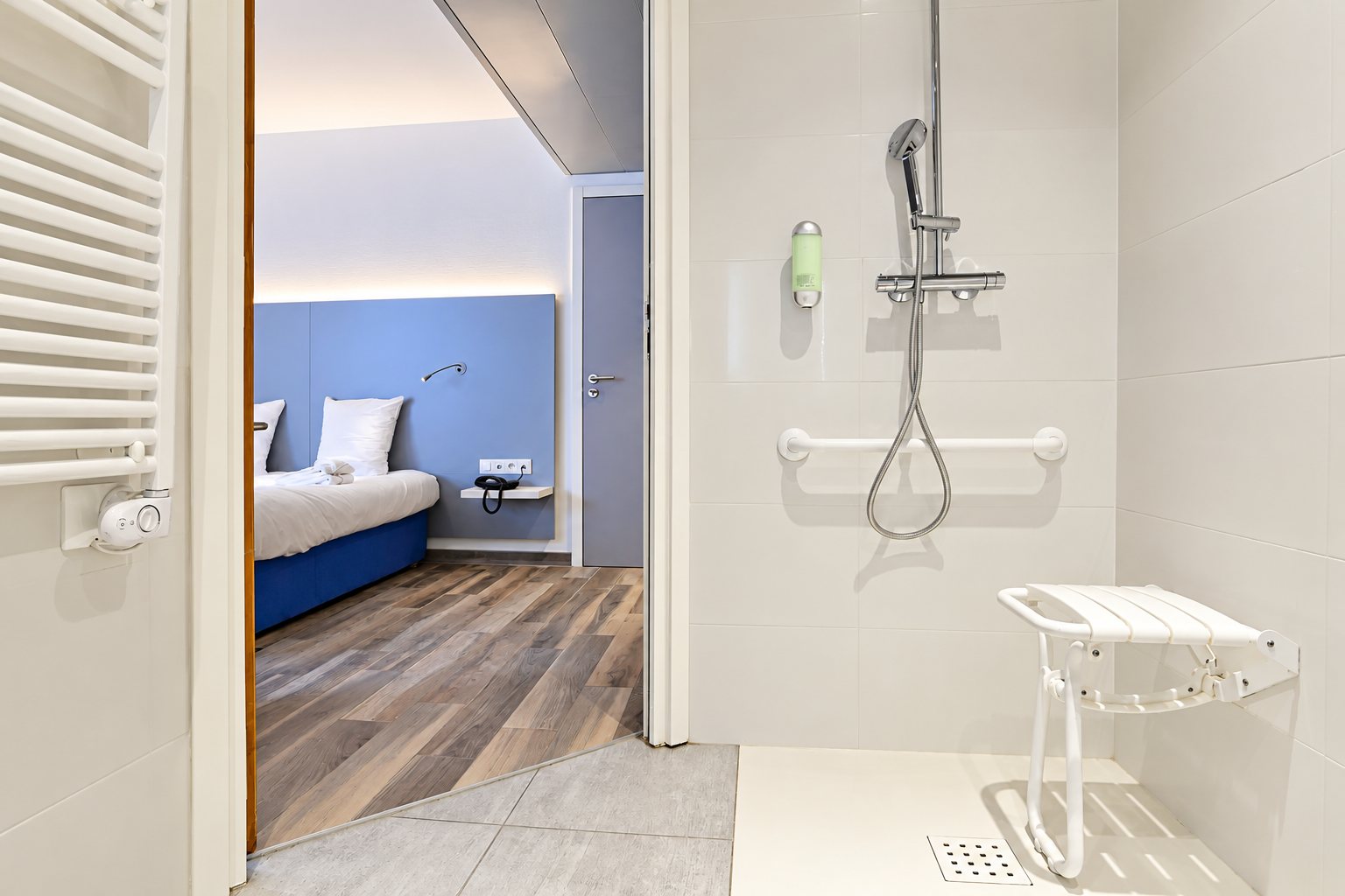 Douche de plain-pied avec barre d'appui et siège rabattable blanc, ouverte sur une chambre d'hôtel aux tons bleus.