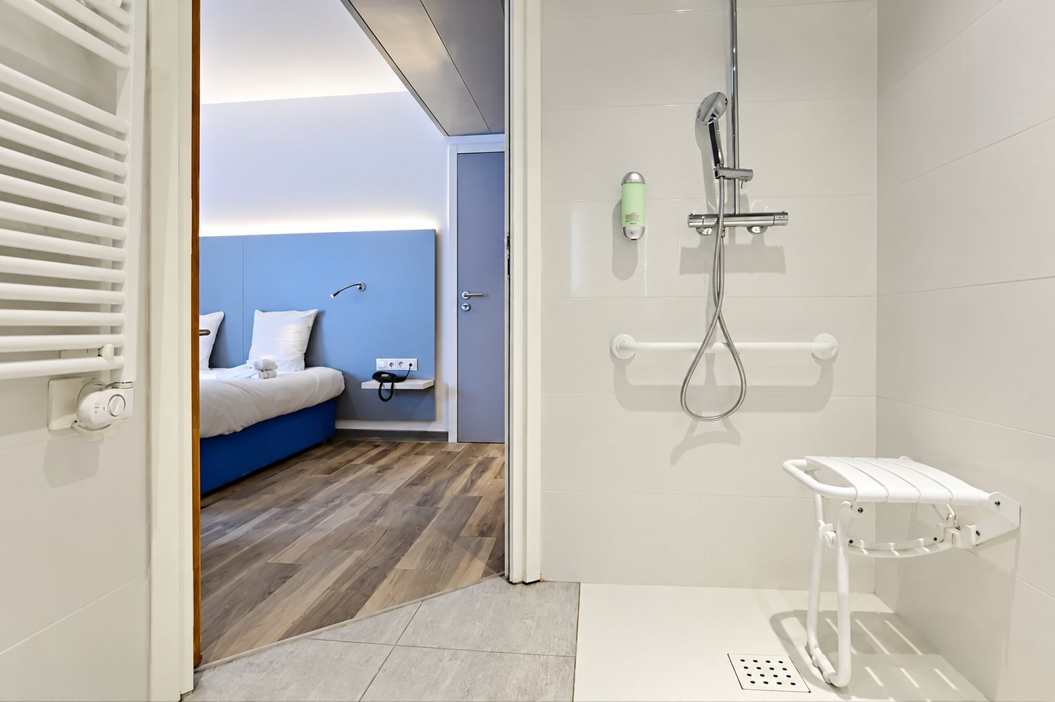 Salle de douche blanche équipée d'une barre d'appui et d'un siège rabattable, ouverte sur une chambre d'hôtel aux tons bleus.