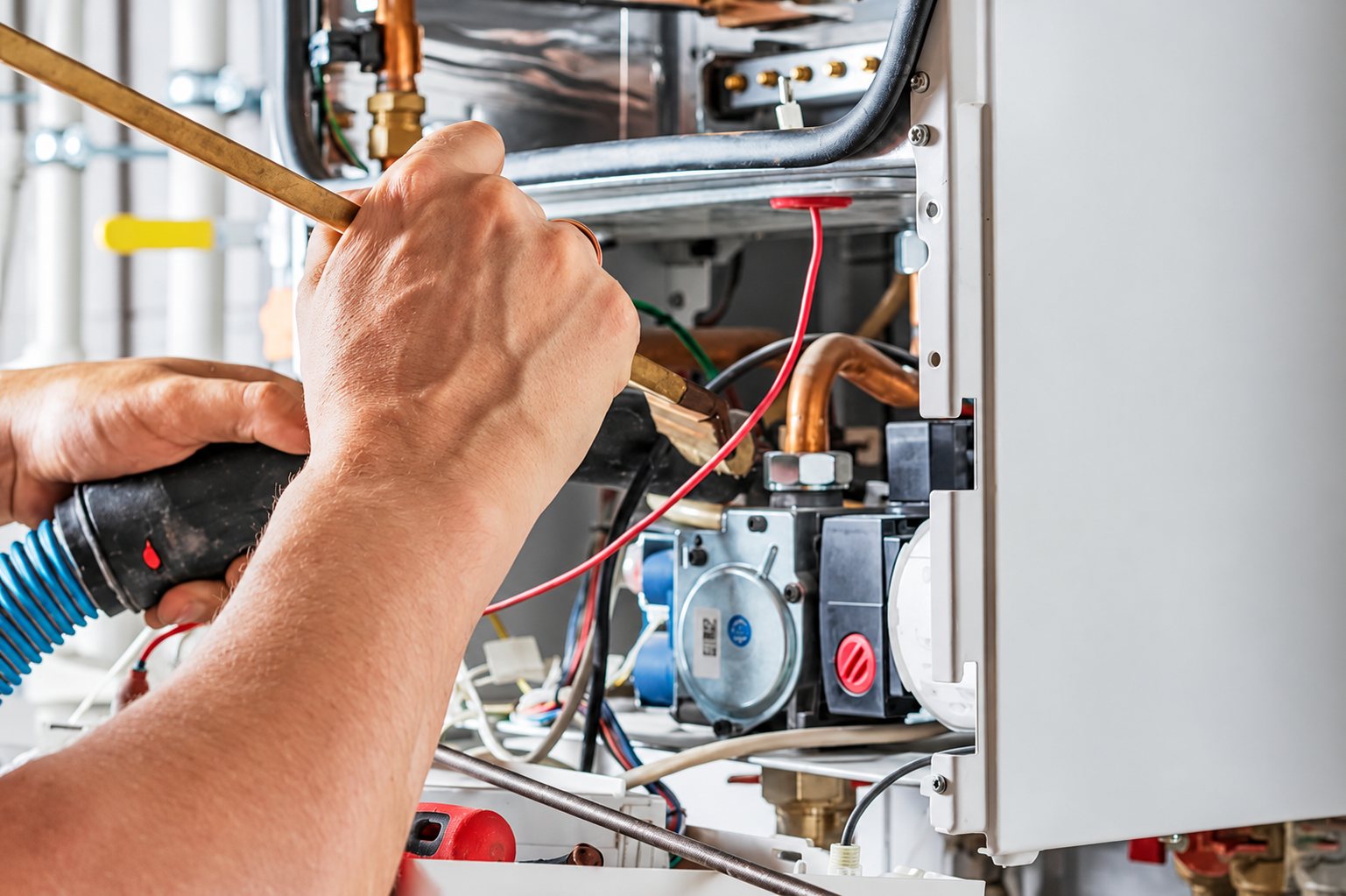 Les mains d'un technicien nettoient l'intérieur d'une chaudière ouverte avec une brosse, près de tuyaux en cuivre et de câbles.