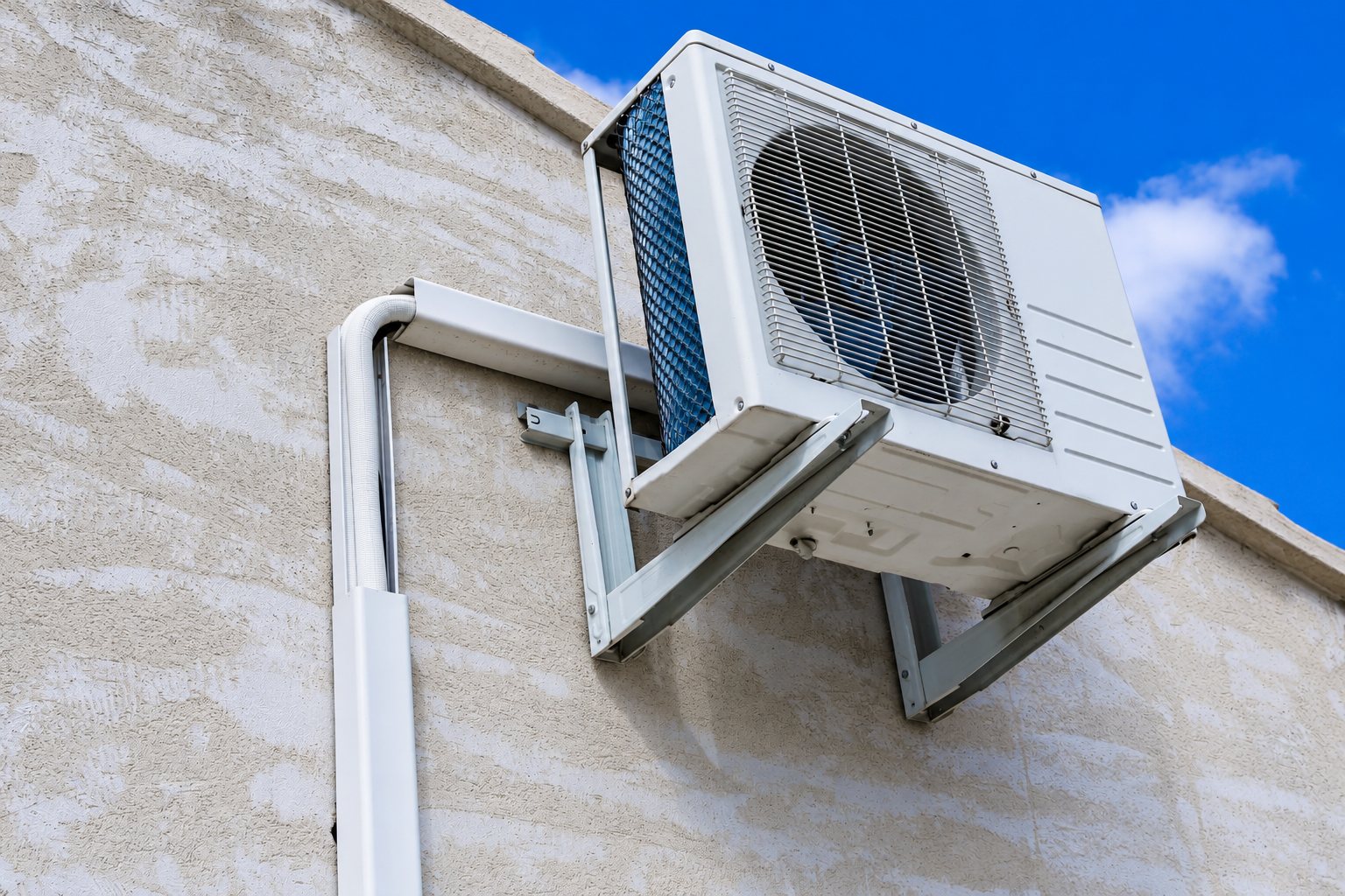 Une unité extérieure de climatisation blanche est installée sur des supports métalliques contre un mur crépi, avec une goulotte blanche et un ciel bleu en arrière-plan.